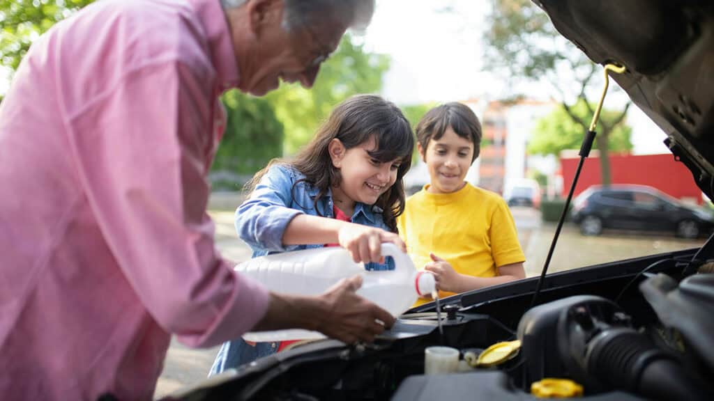 photo d'un homme au dessus d'une voiture avec le capot ouvertet des enfants qui tiennent et versent du liquide de refroidissement dans la voiture - mondial de l'auto 2026